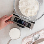 flour in a bowl on a kitchen scale next to measuring cup full with flour and spoon.