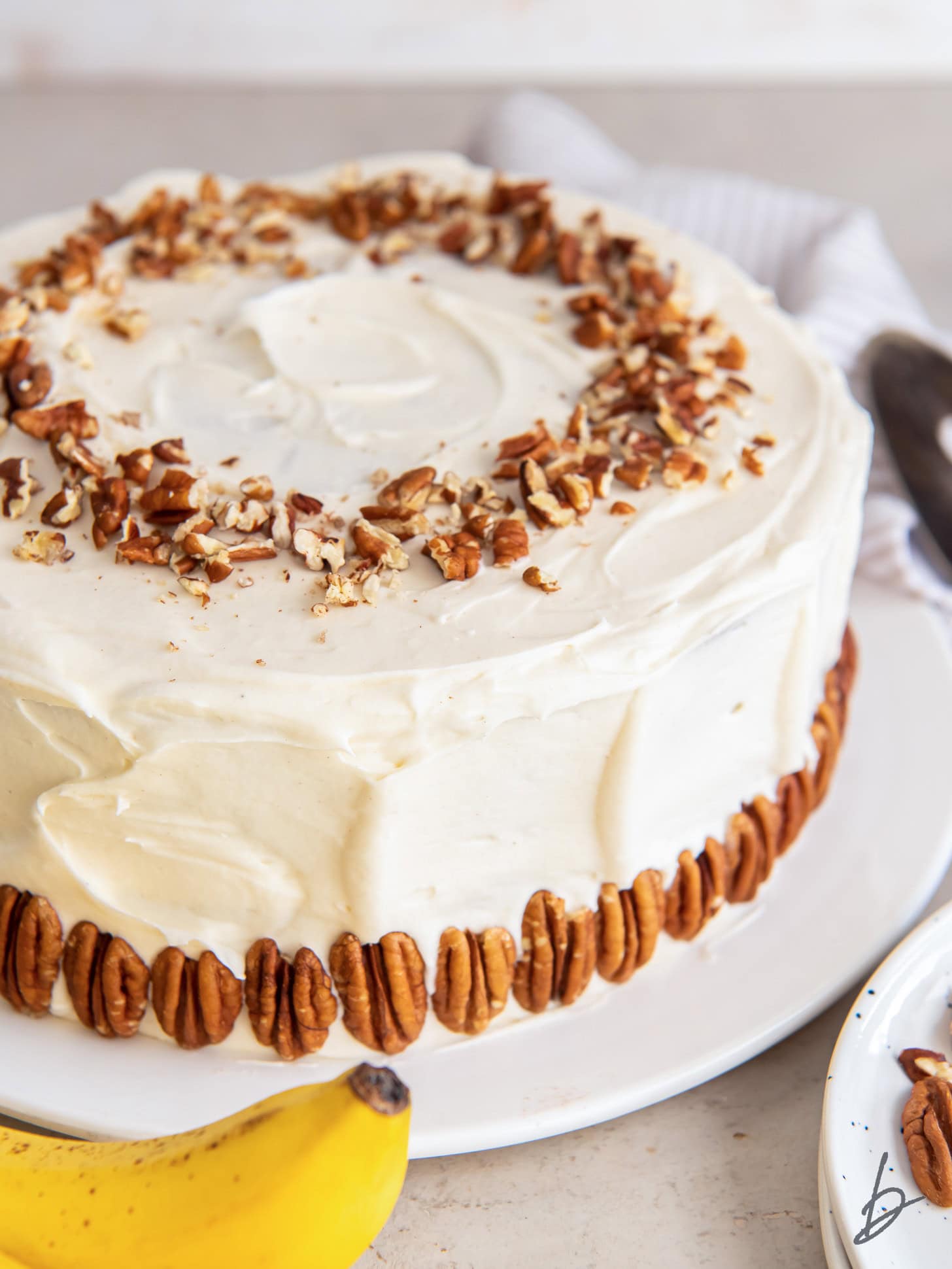 hummingbird cake covered in cream cheese frosting and garnished with pecan halves on outside and chopped pecans sprinkled on top.