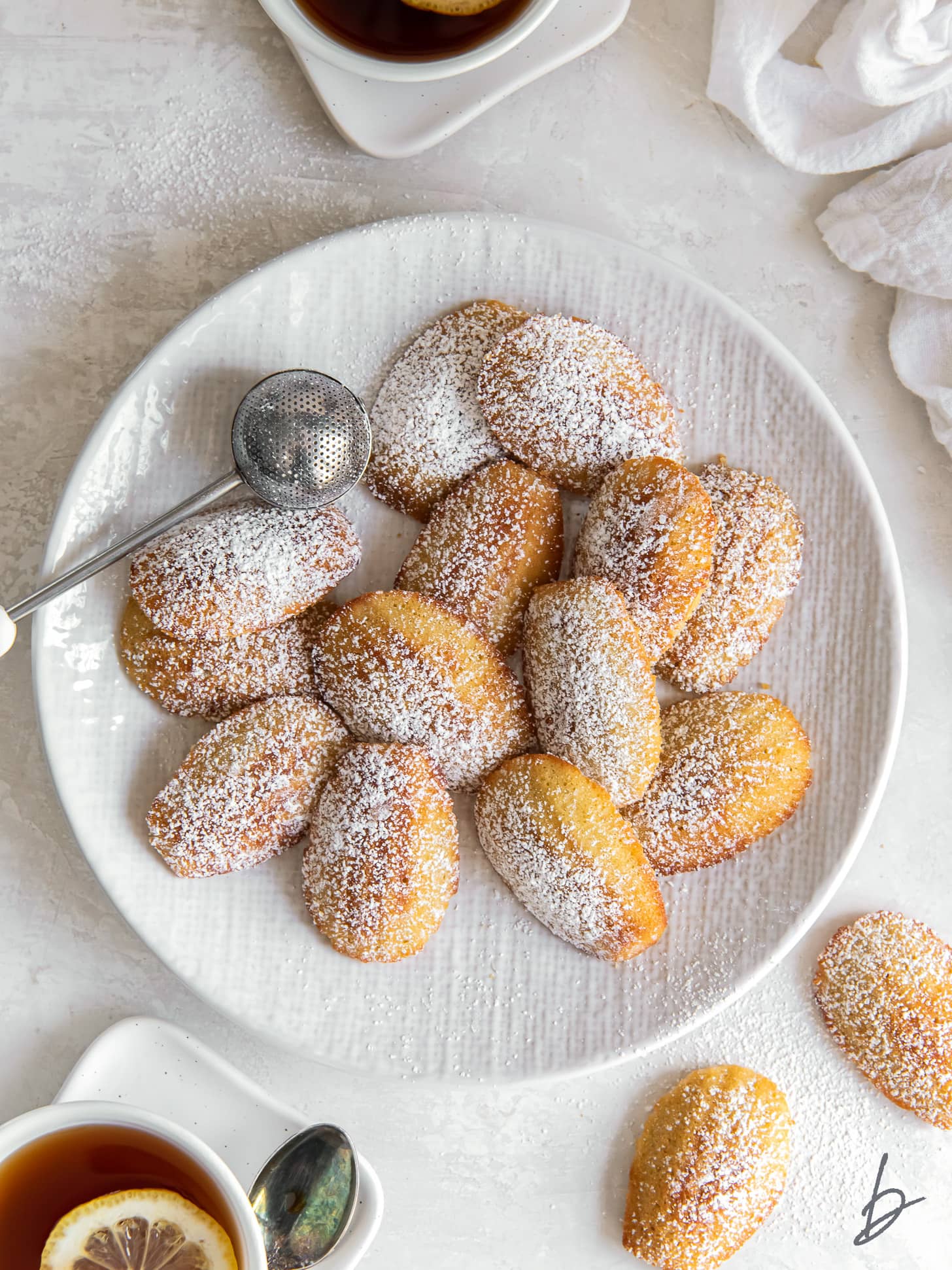 madeleine cookies dusted with confectioners' sugar on a plate next to mug of tea.