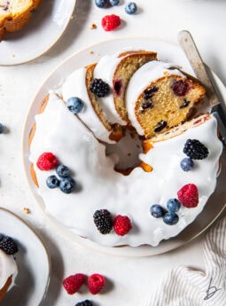 tabletop with a mixed berry pound cake topped with icing and a few slices cut.