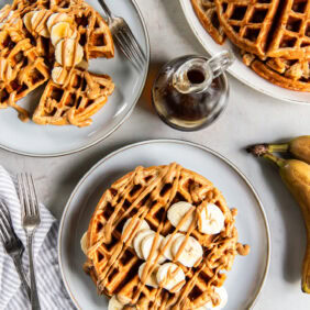 Tabletop with plates of peanut butter banana waffles garnished with fresh banana slices and peanut butter.