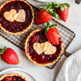 two strawberry jam tarts with shortbread heart cut out and strawberry garnish on small wire cooling rack.