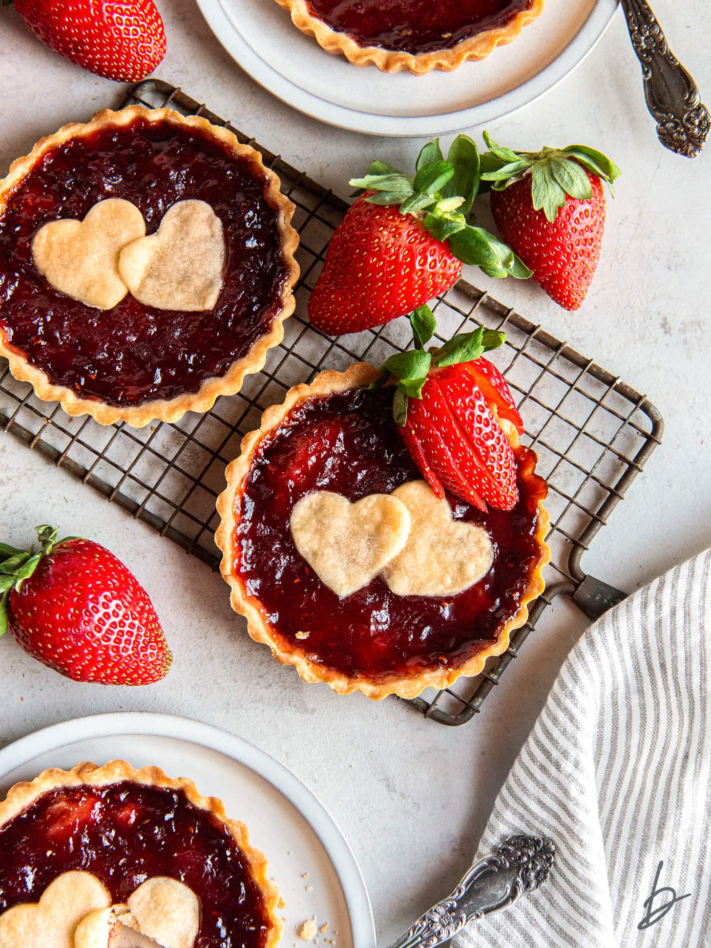 two strawberry jam tarts with shortbread heart cut out and strawberry garnish on small wire cooling rack.