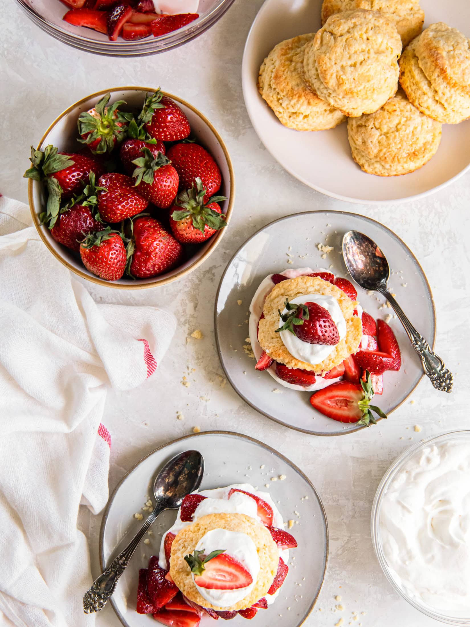 two strawberry shortcakes on plates with spoons next to a bowl of fresh strawberries.