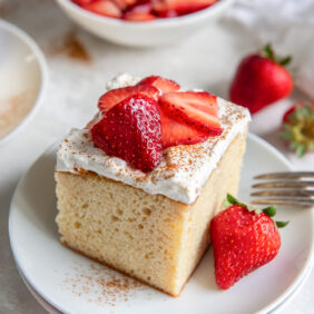 square slice of tres leches cake on a plate with whipped cream topping and strawberries.