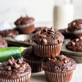 double chocolate zucchini muffins in a stack in front of muffin tin and glass milk bottle.