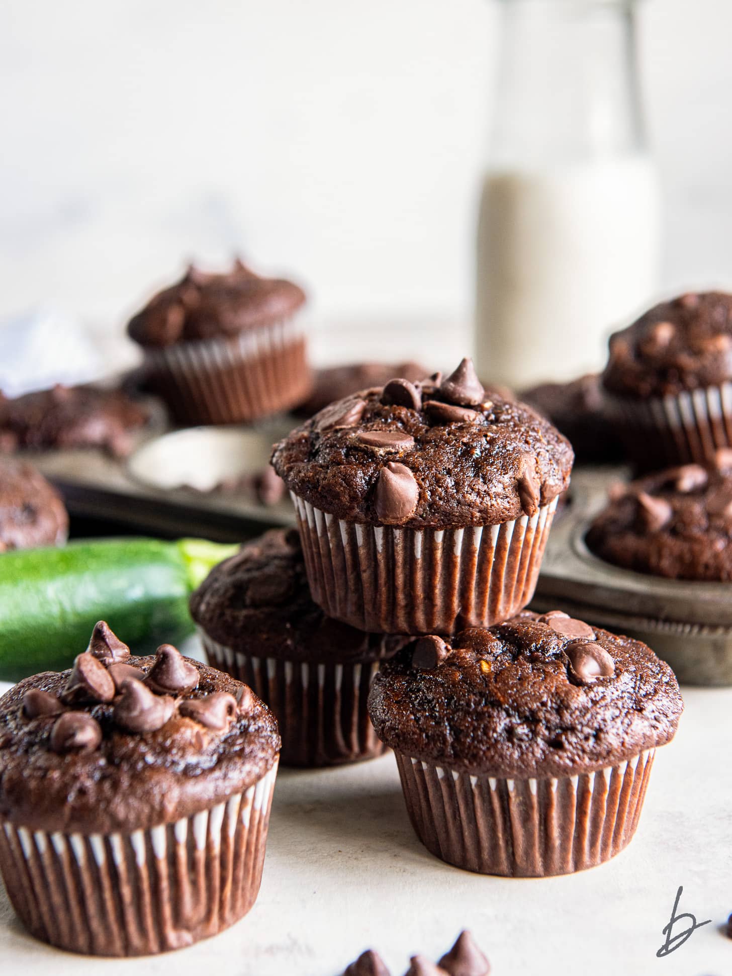 double chocolate zucchini muffins in a stack in front of muffin tin and glass milk bottle.