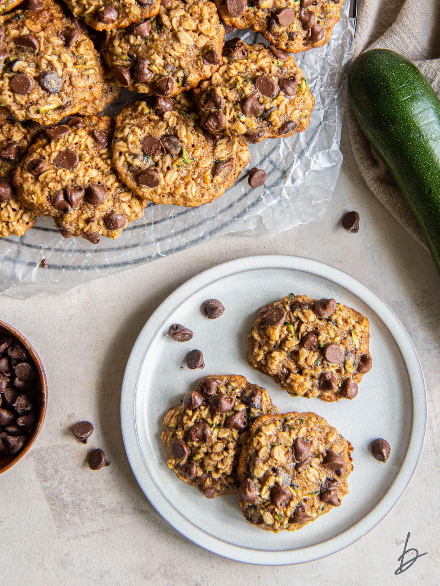 chocolate chip zucchini cookies on a plate next to a zucchini and more cookies on a wire rack.
