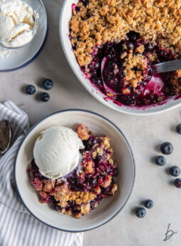 blueberry crisp in a bowl with a scoop of vanilla ice cream next to dish of more fruit crisp.