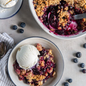 blueberry crisp in a bowl with a scoop of vanilla ice cream next to dish of more fruit crisp.