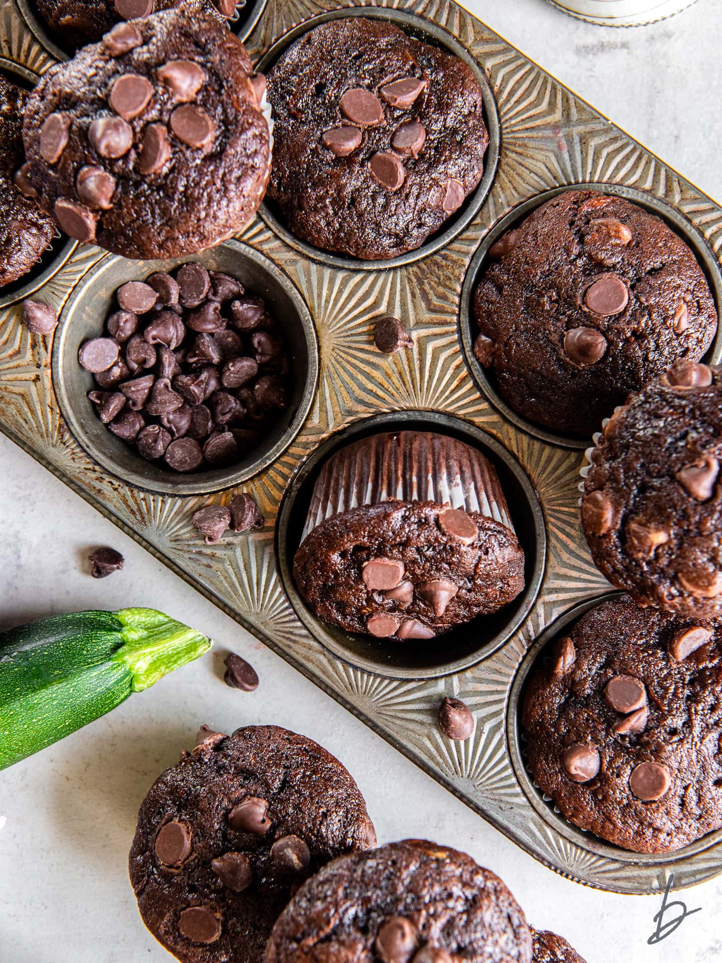 double chocolate zucchini muffins in a muffin tine with chocolate chips and zucchini next to it.