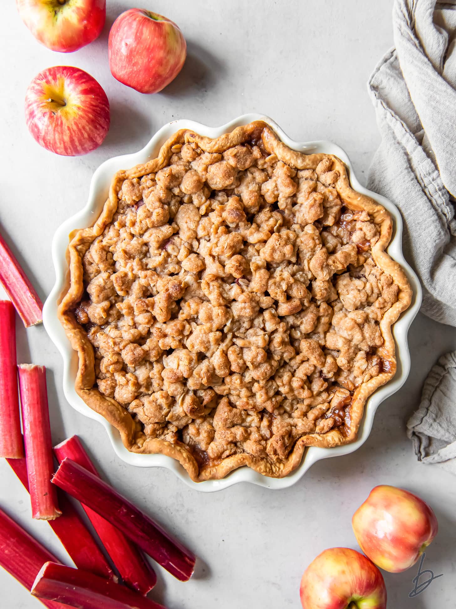 unsliced apple rhubarb pie with crumb topping next to fresh apples and rhubarb.