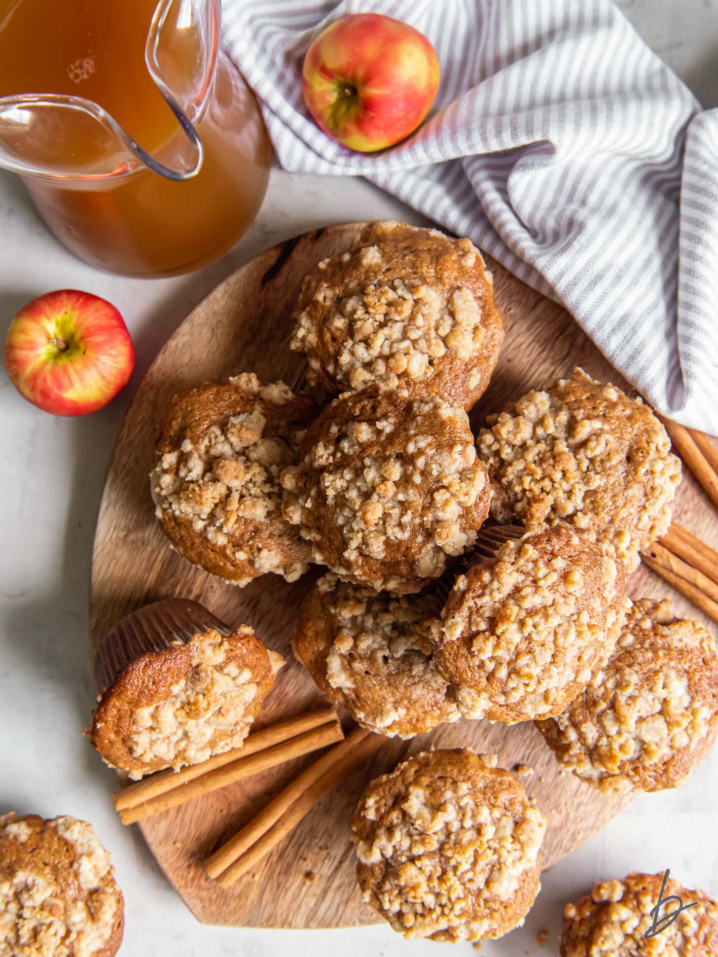 apple cider muffins with streusel topping on a wood platter next to pitcher of cider.