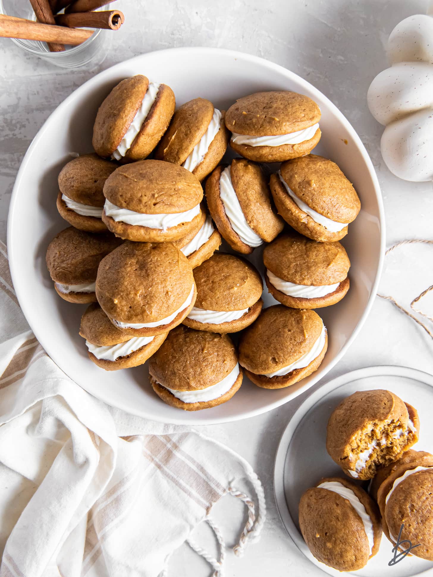 shallow bowl of pumpkin whoopie pies next to white kitchen cloth and jar of cinnamon sticks.