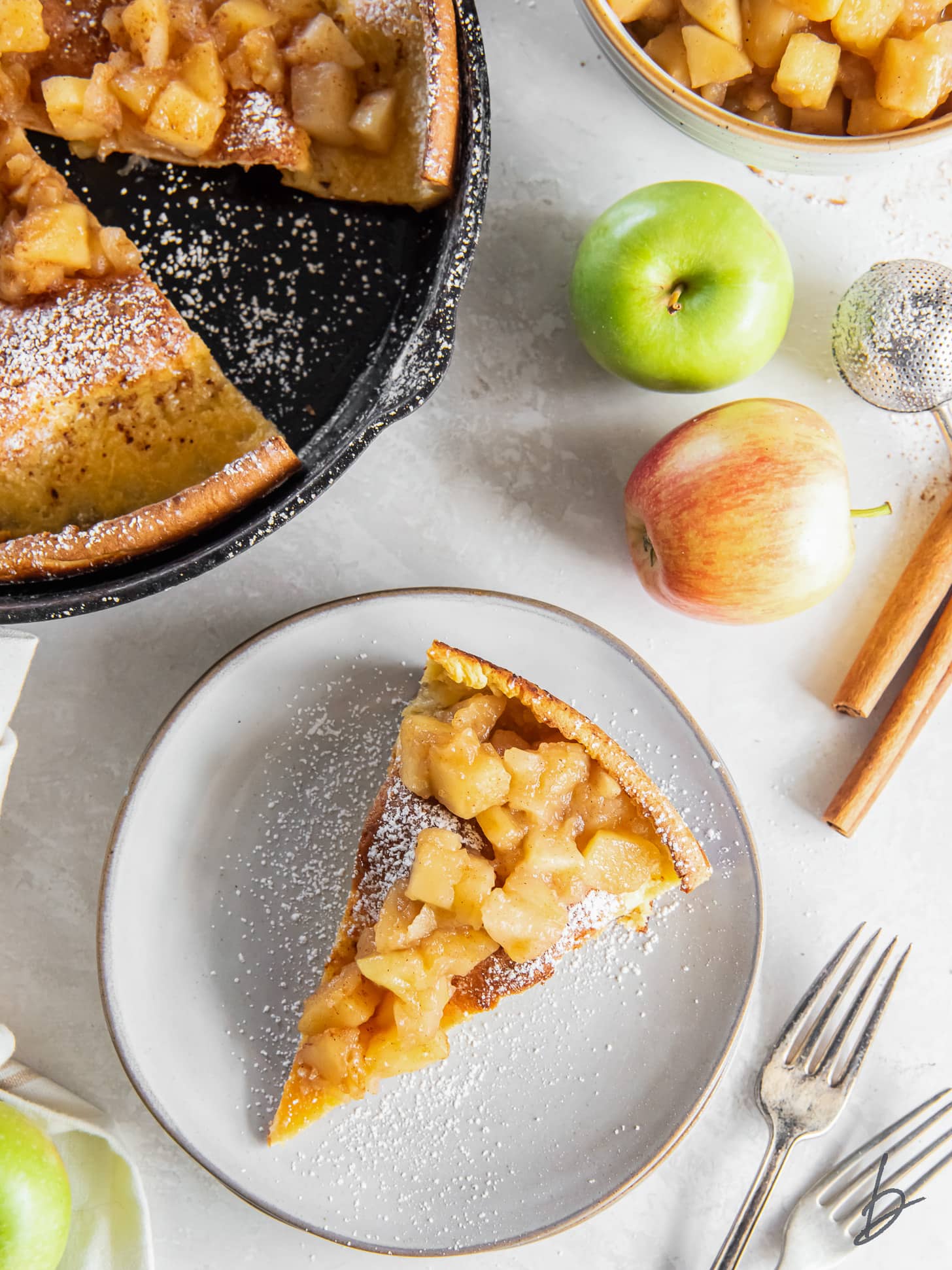 slice of apple dutch baby on a plate next to fresh apples and cinnamon sticks.