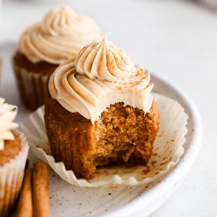pumpkin cupcake with a bite sitting on open paper liner on plate with cinnamon sticks.
