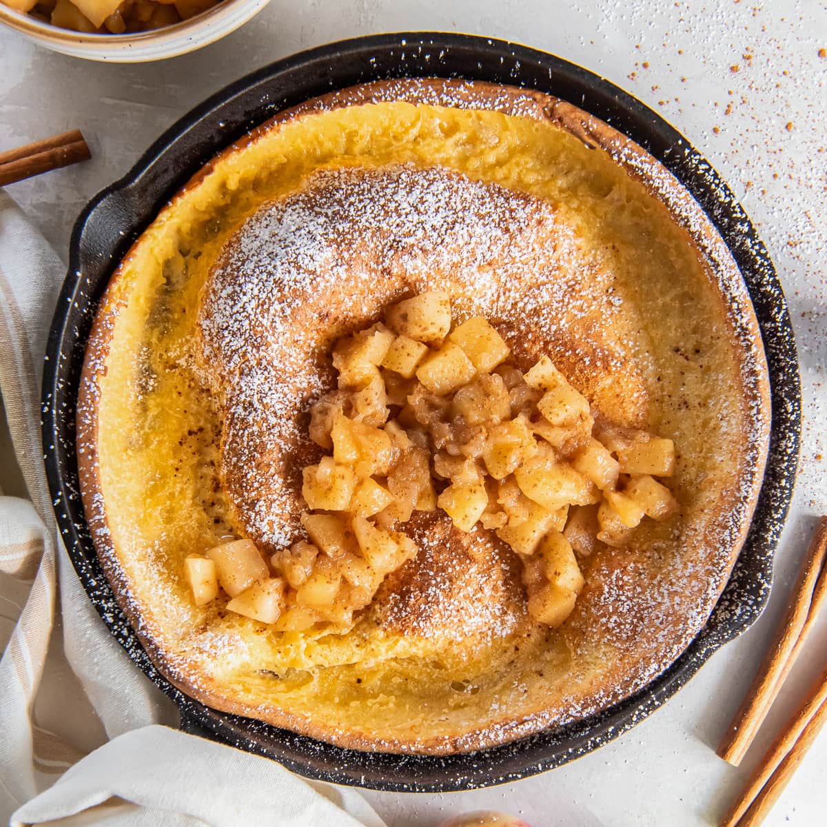 apple dutch baby topped with apple compote and dusted with confectioners sugar in a skillet.