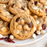 thick and chewy white chocolate cranberry cookie leaning on more cookies.
