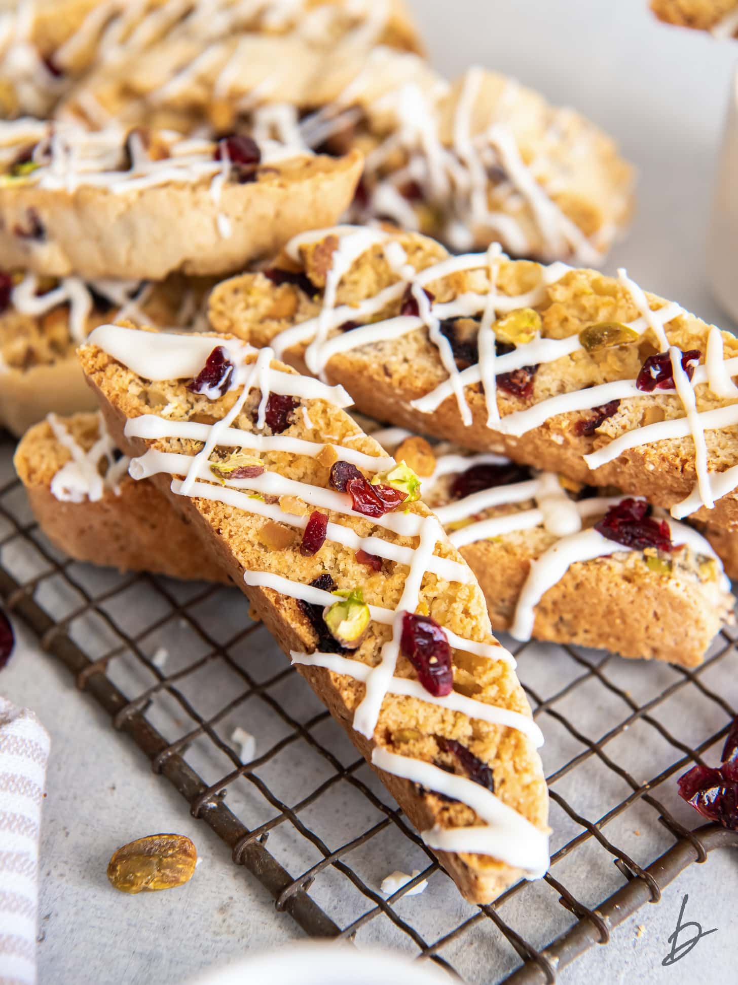 cranberry pistachio biscotti drizzled with white chocolate in a pile on a wire rack.