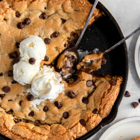 two spoons holding bites in the center of a gooey chocolate chip skillet cookie with vanilla ice cream.