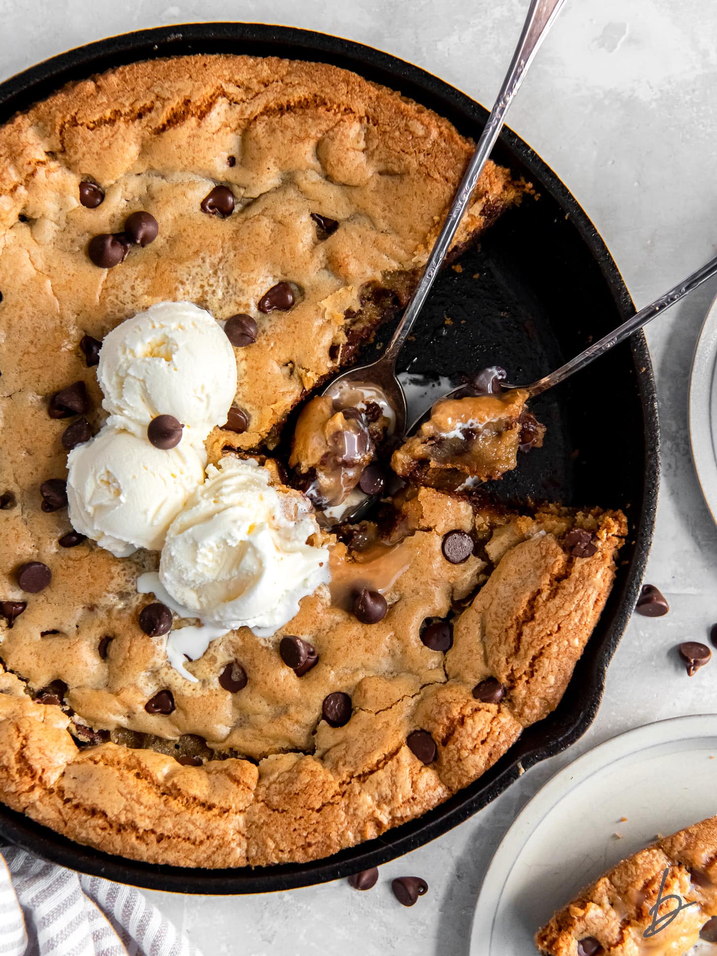 two spoons holding bites in the center of a gooey chocolate chip skillet cookie with vanilla ice cream.