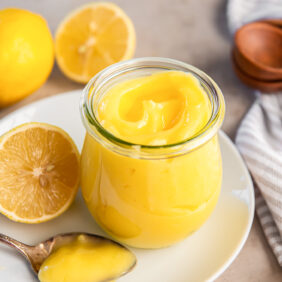 glass jar of lemon curd on a plate next to a lemon half and spoonful of curd.