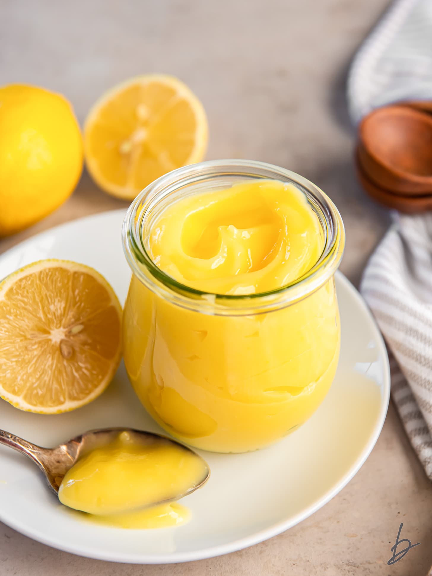 glass jar of lemon curd on a plate next to a lemon half and spoonful of curd.