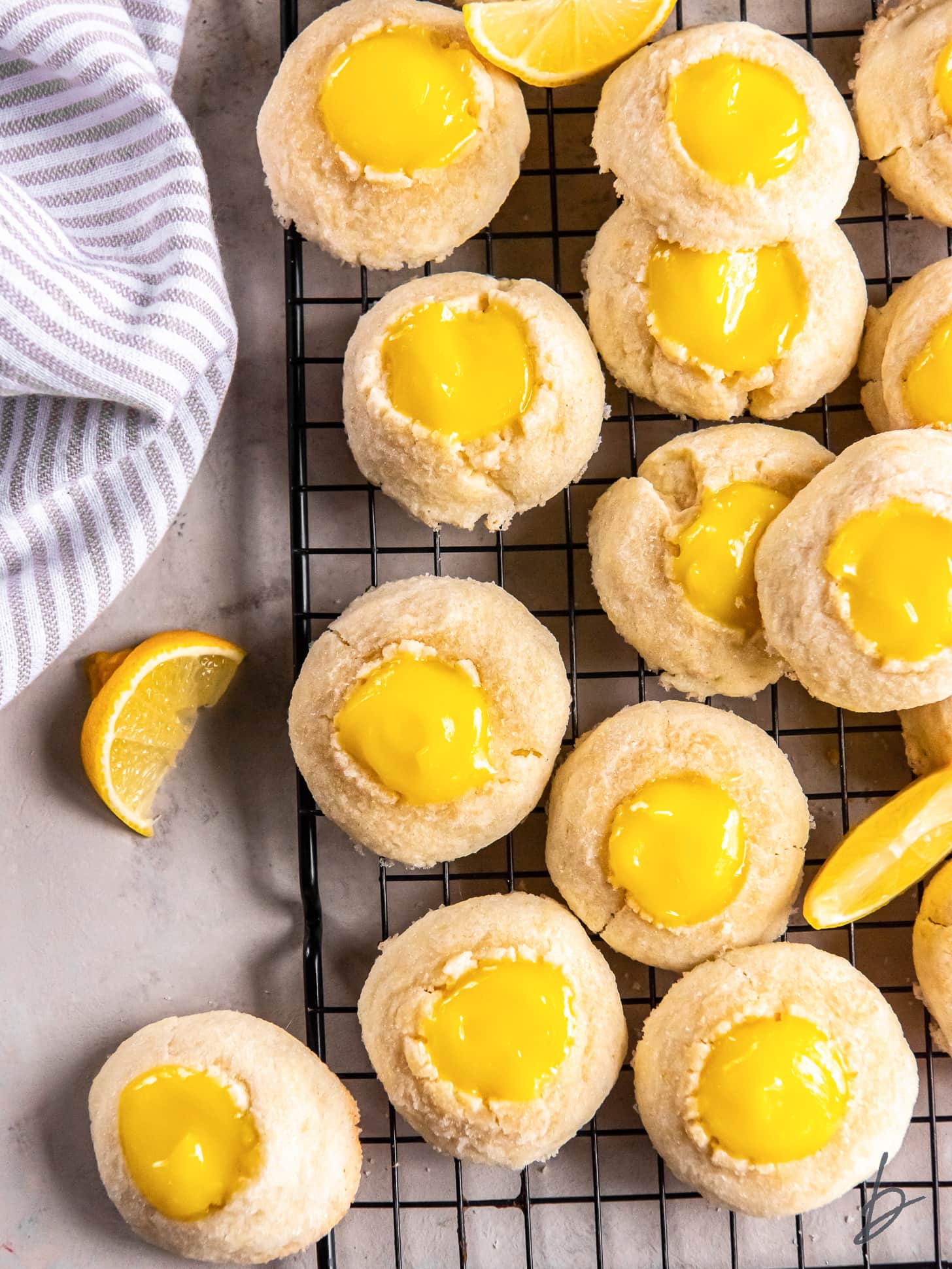 lemon curd thumbprint cookies on a wire rack next to a striped kitchen cloth and lemon wedge.