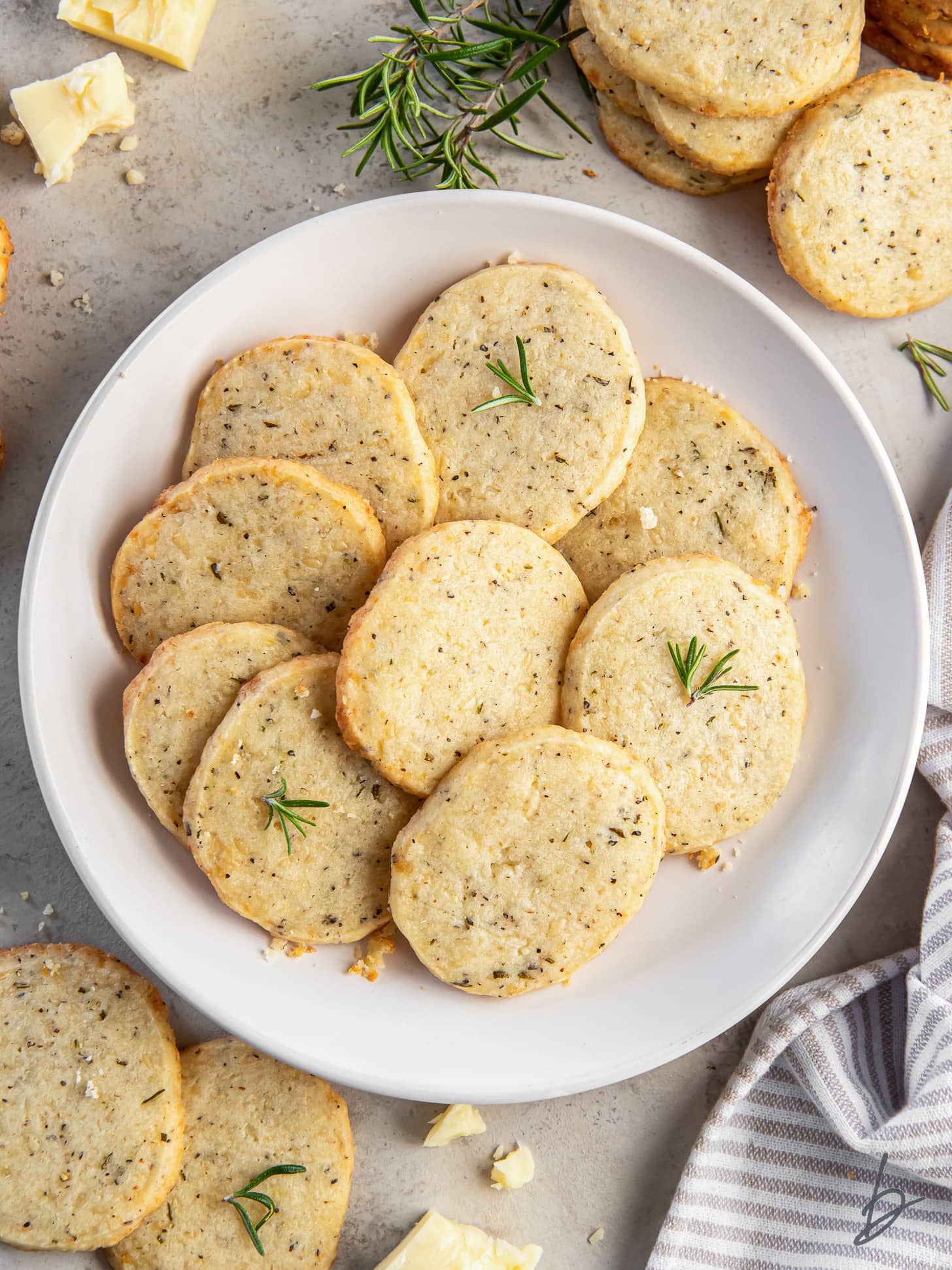 plate of savory shortbread cookies next to a couple crumbles of white cheddar cheese and sprigs of rosemary.