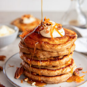 maple syrup poured on top and drizzling down a stack of carrot cake pancakes on a plate.