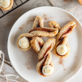 three easter bunny puff pastry twists with cream cheese frosting bunny tails on a white plate.