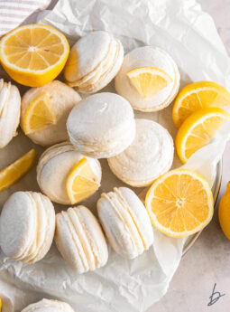 lemon macarons in a pile on parchment paper with lemon wedges.