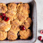Cherry cobbler with biscuit topping in a square baking dish next to a few fresh cherries.