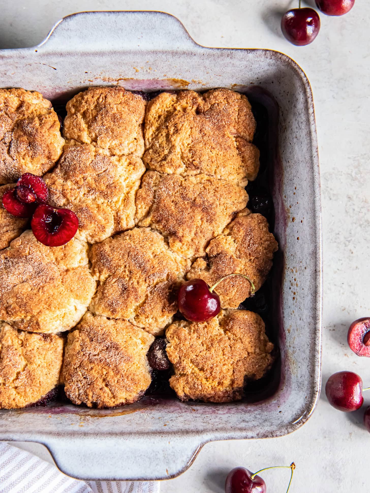 Cherry cobbler with biscuit topping in a square baking dish next to a few fresh cherries.