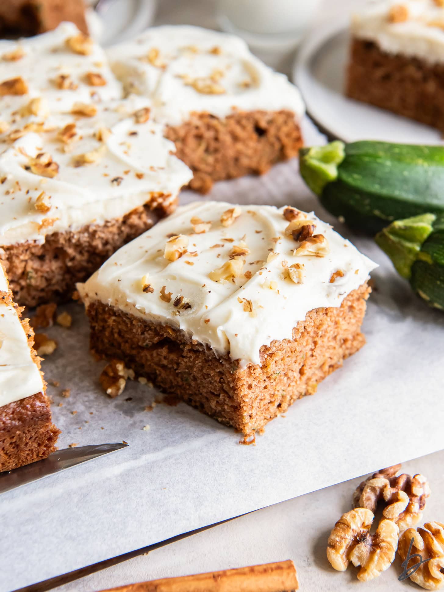 square slice of zucchini spice cake with cream cheese frosting next to fresh zucchini and more cake.