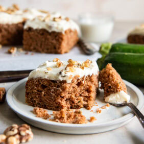 square slice of zucchini spice cake on a plate with a bite taken out of it by a fork.