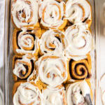 13x9 baking dish with pumpkin spice cinnamon rolls being frosted with cream cheese icing.
