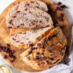 tabletop with circle cutting board and a loaf of no knead cranberry bread on top of it with a few slices cut.