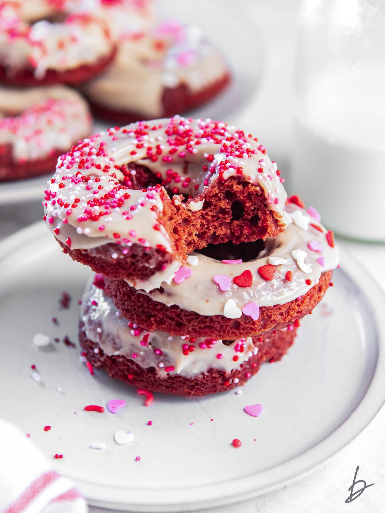stack of three red velvet donuts with cream cheese icing and sprinkles; top donut with a bite.