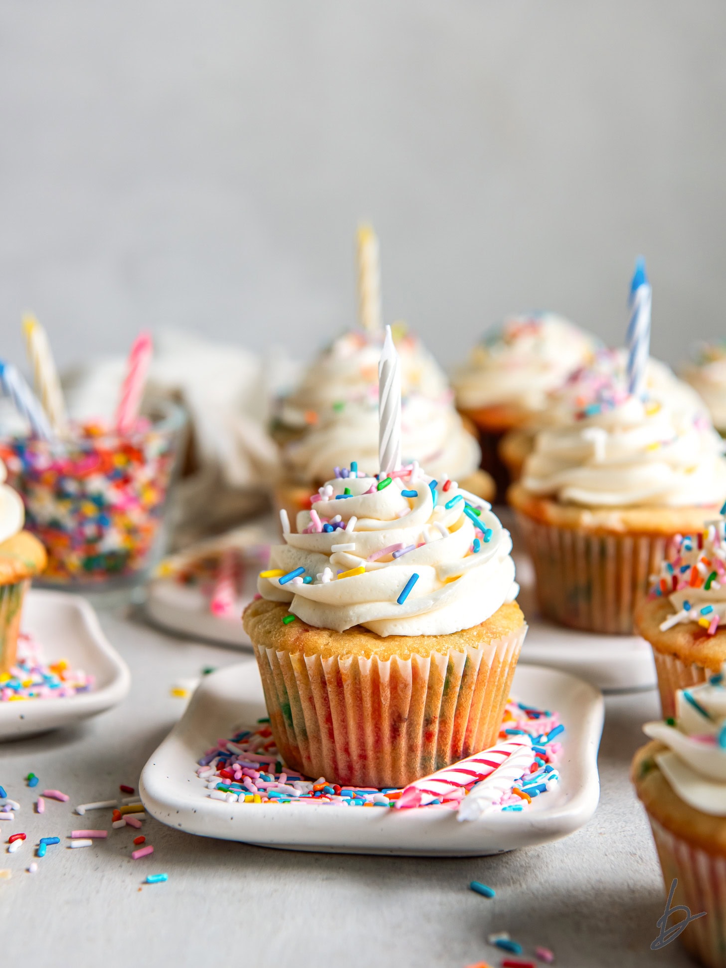 plate with a funfetti cupcake with sprinkles and a birthday candle in front of more cupcakes on a tabletop.