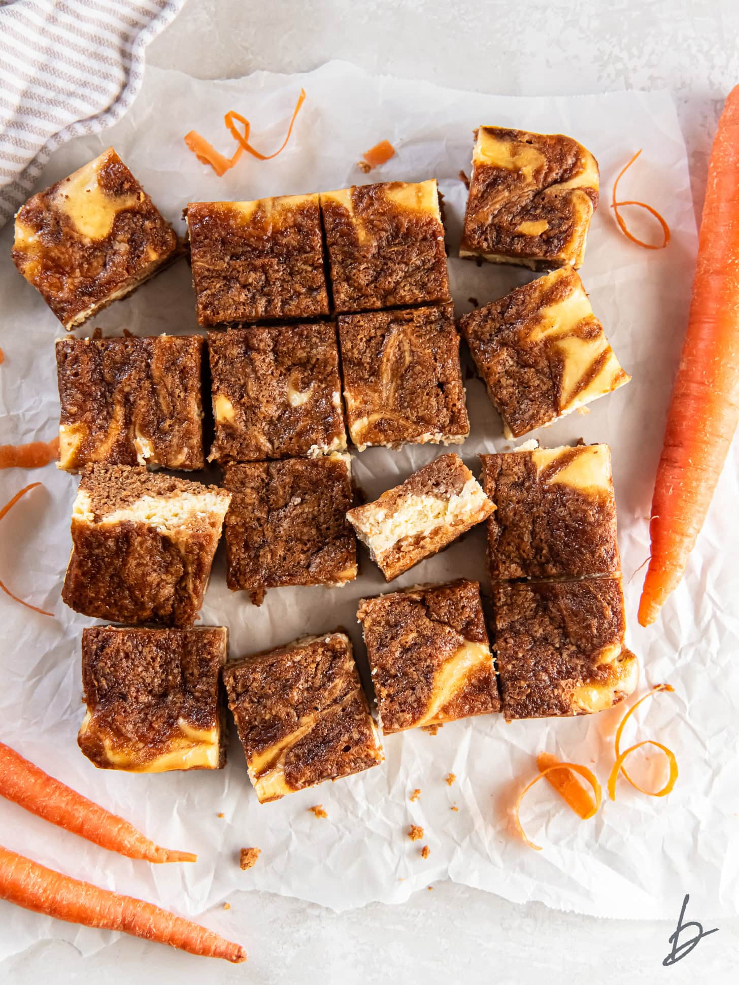 Bars with layers of cheesecake and carrot cake on parchment paper near carrots.