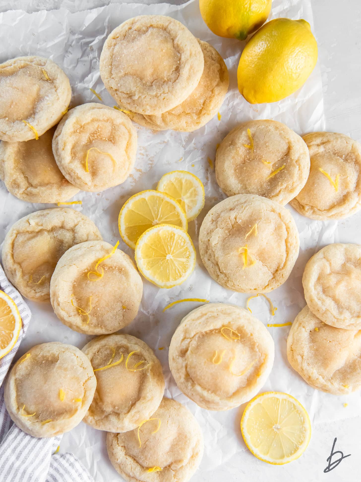 tabletop with parchment paper and lemon cheesecake cookies scattered on top with lemon slices.