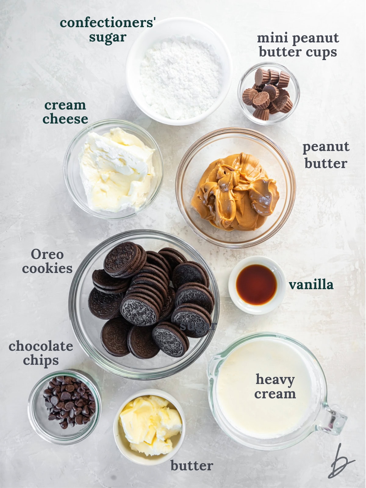 A bowl with Oreo cookies near other small bowls with peanut butter, candies, cream cheese, heavy cream and butter.