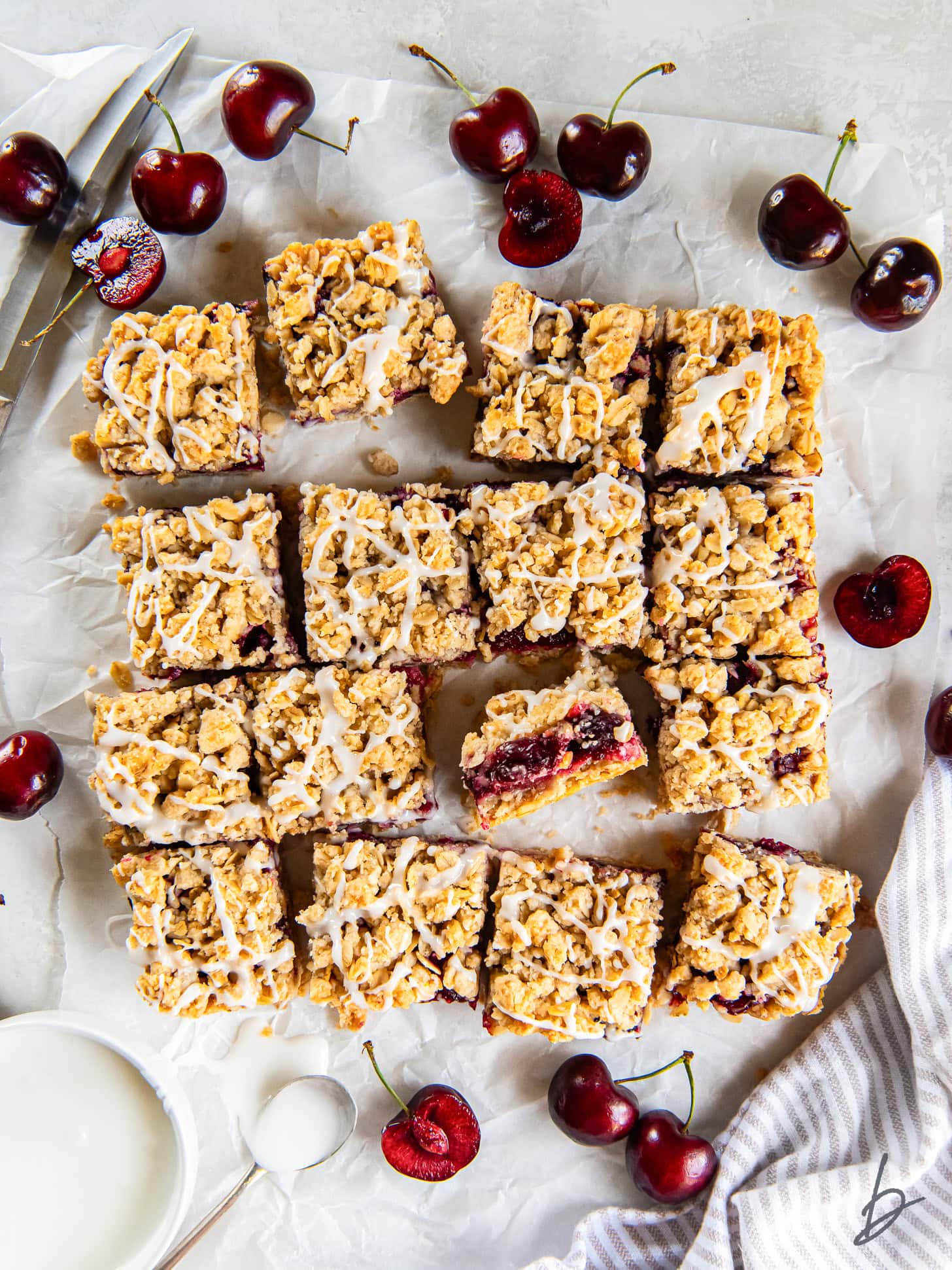 tabletop with cherry crumble bars with almond icing on parchment paper next to fresh cherries.