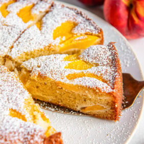 cake server holding slice of peach cake on a cake plate with the whole cake dusted in confectioners' sugar.