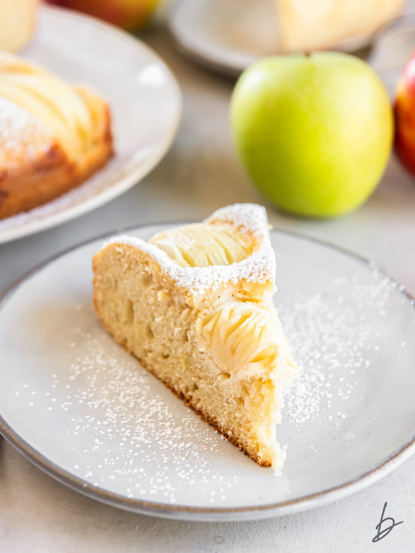 Slice of apple cake on a white plate with a Granny Smith apple in the background.