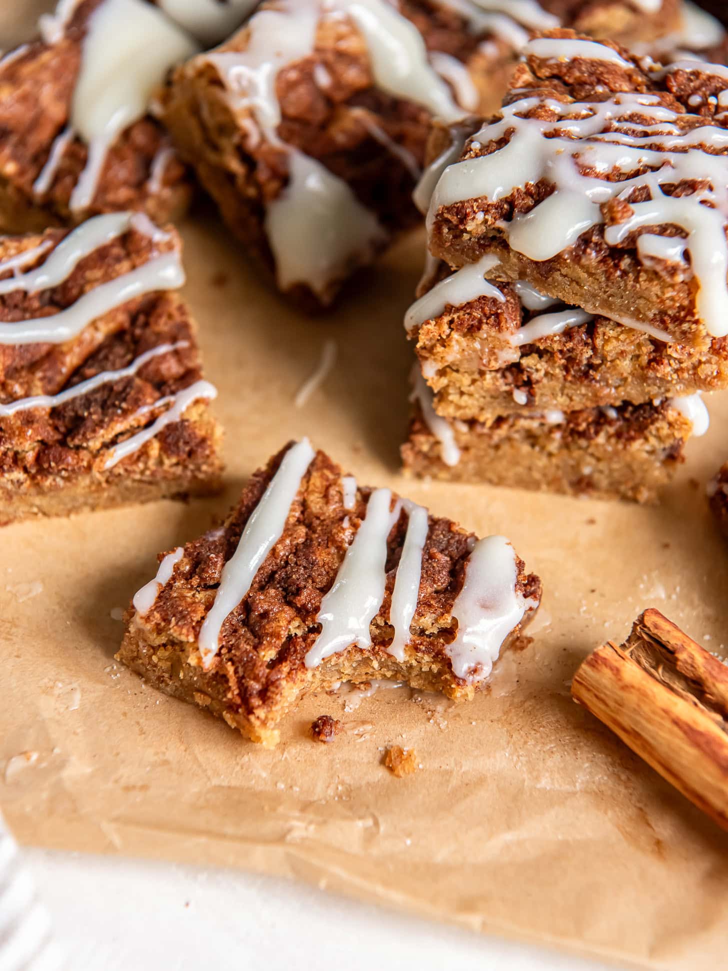 cinnamon roll blondie with a bite taken out of it sitting on parchment paper next to a cinnamon stick.