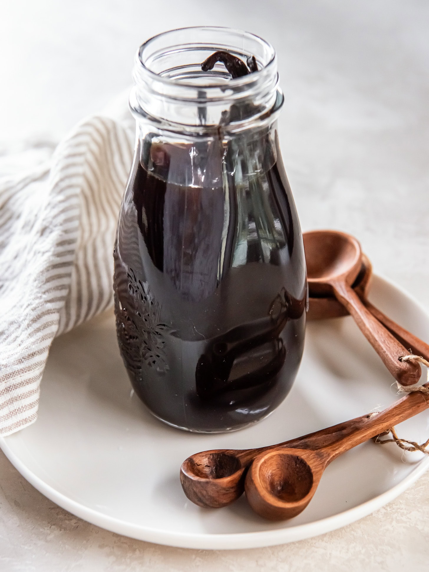 Homemade vanilla extract in a small jar sitting on a white plate near small wooden spoons.