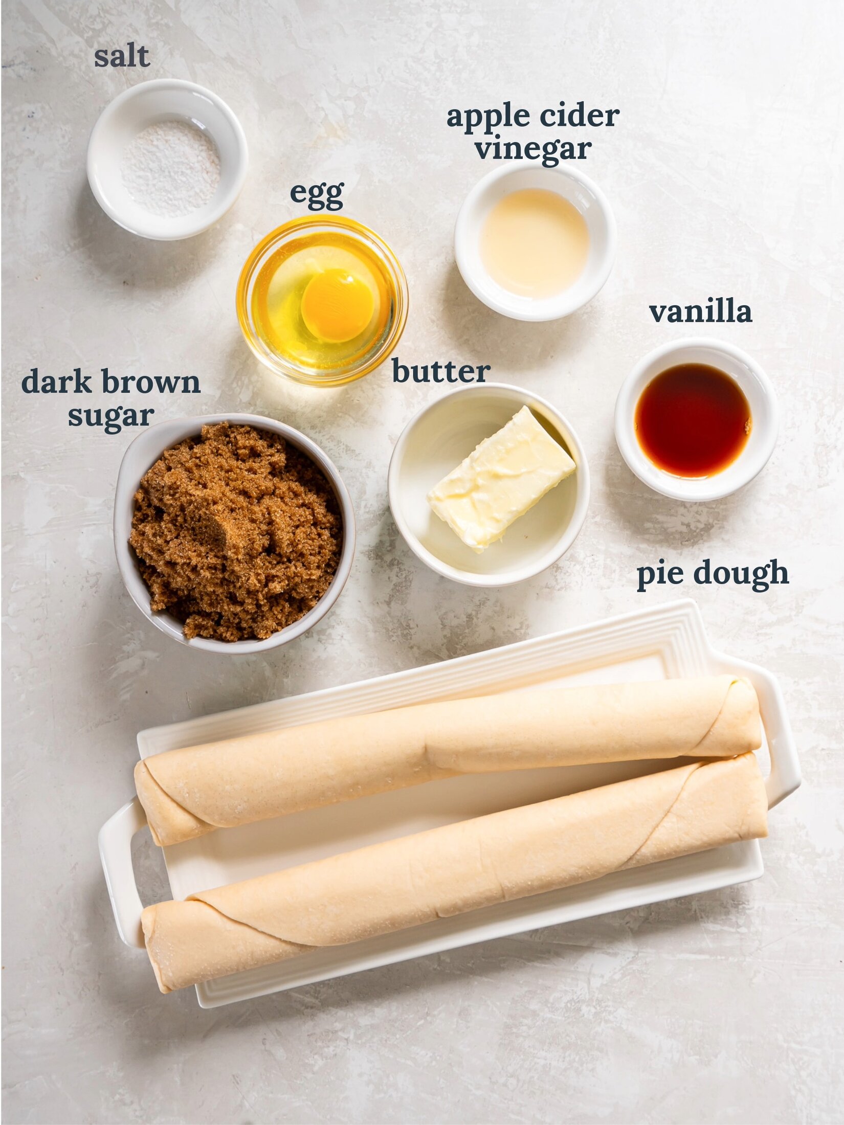 Two rolls of pie dough near small bowls with brown sugar, vanilla, egg, and butter.