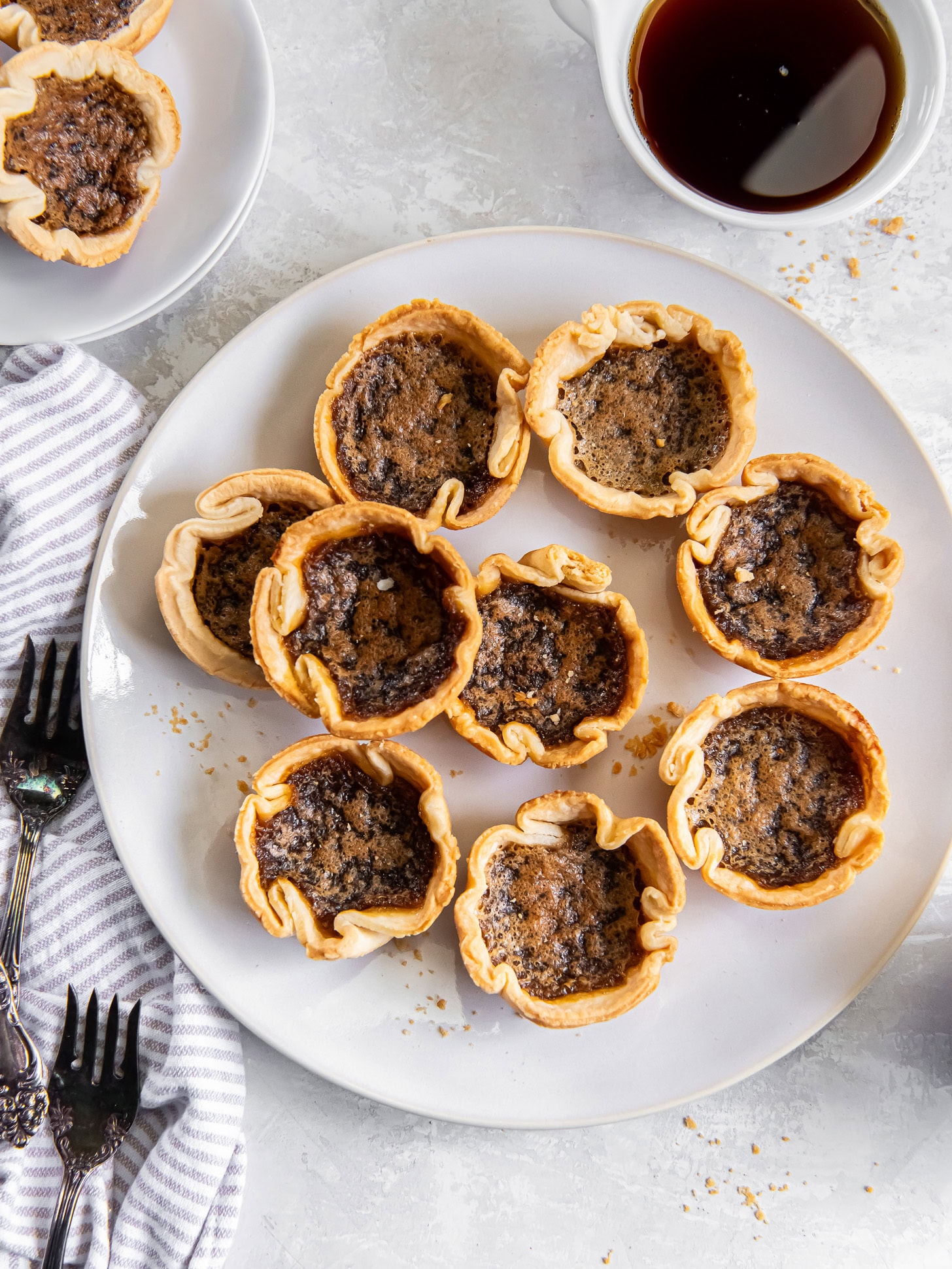 Butter tarts on a large white plate near forks and white mug of coffee.